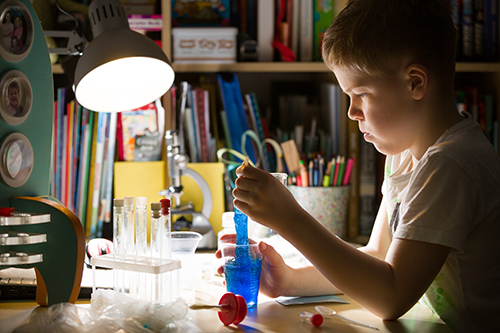 ילד בבית ספר יסודי חמוד בודקים מיקרוסקופ ליד שולחנו בבית. מדען צעיר עושה ניסויים במעבדה בביתו. בתוך בית. ילד ומדע.